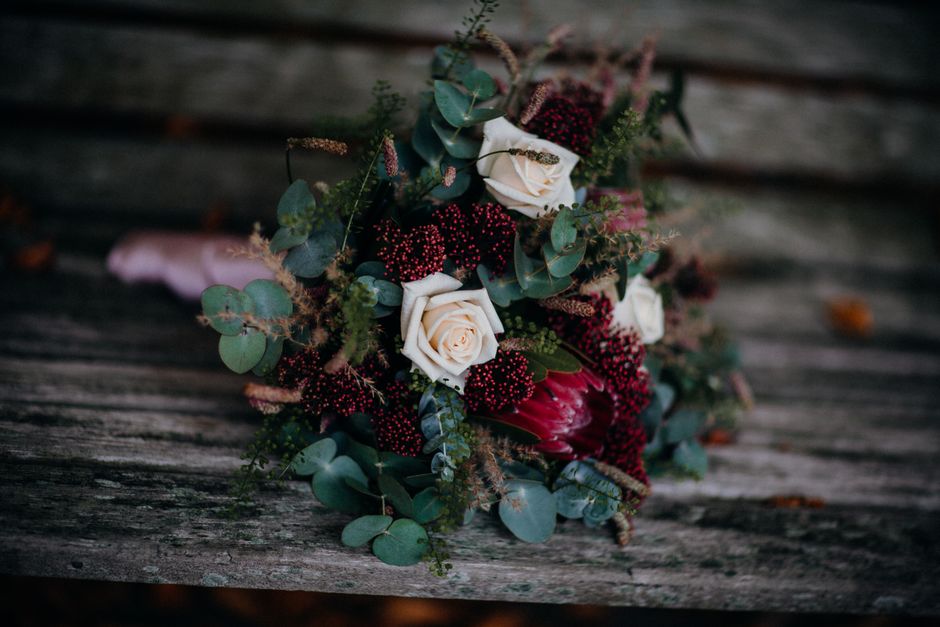 A bouquet featuring two white roses surrounded by clusters of small, deep red berries and sprigs of eucalyptus with round, bluish-green leaves. The flowers are arranged on a rustic wooden surface with