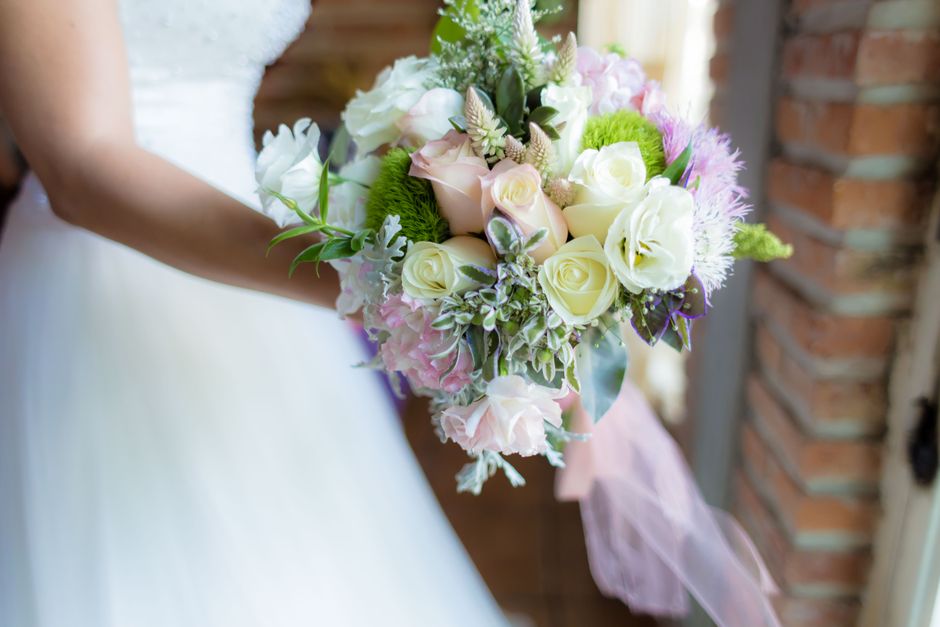 A bride dressed in a white wedding gown is holding a round bouquet of fresh flowers, featuring white roses, pale pink lisianthus, and light purple blooms, accented with green foliage and variegated le