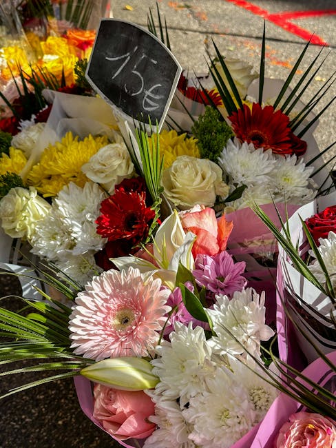 An assortment of fresh floral bouquets arranged outdoors, featuring pink gerbera daisies, white roses, red gerbera daisies, and creamy white chrysanthemums, with green palm leaves and long, slender gr