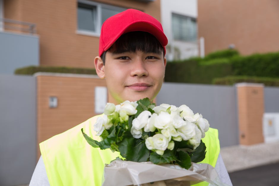 A young man wearing a red cap and a high-visibility yellow jacket holds a bouquet of fresh white roses wrapped in light brown paper. He is standing outdoors in front of modern residential buildings wi