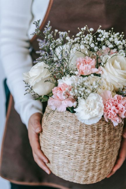 A person dressed in a white shirt and brown apron holds a woven wicker basket filled with a fresh floral arrangement. The bouquet features soft pink carnations, white roses, and sprigs of baby's breat