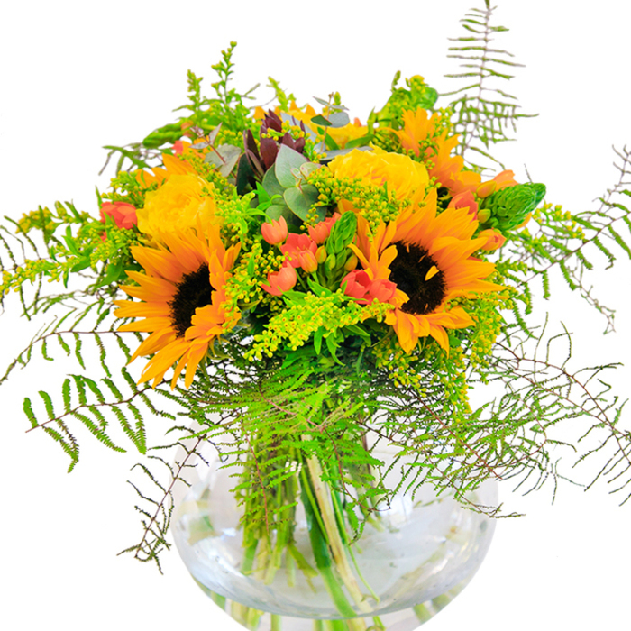Full of optimism and artful flair, this bouquet by Winchmore Hill Florist is a tribute to the golden light that spills onto Winchmore Hill's vibrant streets and friendly parks. The composition revolves around a cluster of striking sunflowers, their wide-open, sunflower-yellow petals and dark, textured faces creating a dramatic yet cheerful focal point-a nod to the communal warmth felt on The Green during local celebrations. Surrounding them are creamy yellow roses, each petal softly curved and layered, radiating classic elegance and a gentle, honeyed aroma. Lively bunches of rich red Hypericum berries add a spirited accent, reminiscent of the bursts of colour often spotted in local gardens along Grovelands Park. Ferny foliage, slender spikes of golden filler, and subtle blue-green eucalyptus sprigs thread their way through the bouquet, imparting gentle movement and cooling verdancy. The arrangement rests in a pristine, round glass vase, whose transparent body reveals the interplay of robust green stems and glistening water-suggesting both vitality and craftsmanship. Sunlight dances across the flowers, accentuating their vivid hues and casting delicate shadows that bring a sense of depth and calm. The bouquet's soft, layered fragrances conjure up the freshness of a Winchmore Hill morning. A perfect gesture for anniversaries, special days, or simply sharing a piece of local beauty, it exudes happiness and the closeness of a well-loved neighbourhood.